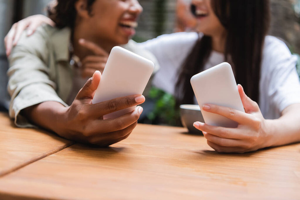 Cropped view of blurred interracial girlfriends holding smartphones in outdoor cafe  - Foto, Imagen