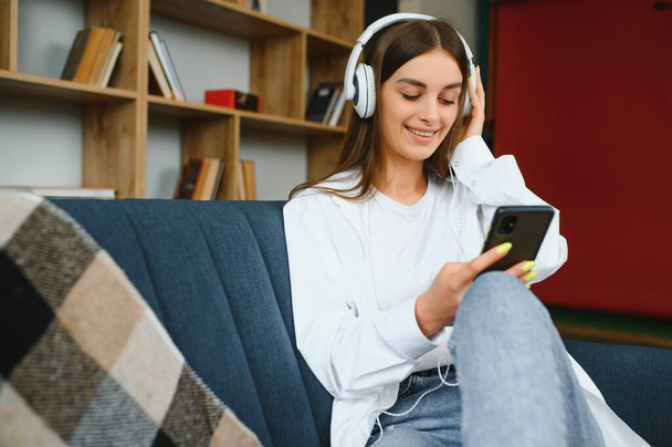 Portrait of attractive girl 20s wearing headphones using smartphone and listening to music while sitting on sofa at home. - Φωτογραφία, εικόνα