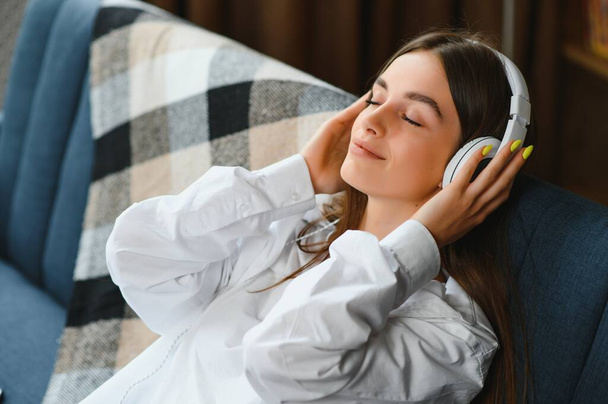 Beautiful young caucasian woman in casual clothing enjoying music and smiling while resting at home. Young woman with headphones using laptop at home. - Zdjęcie, obraz
