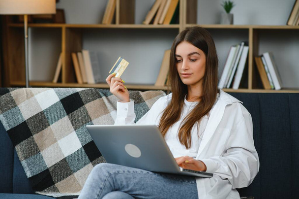 Online Store. Happy girl doing shopping via internet using laptop and credit card, sitting on couch, copyspace. - Photo, image