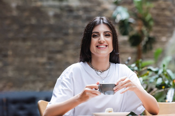 Smiling woman holding cup of coffee near dessert on terrace of cafe  - Foto, Imagen