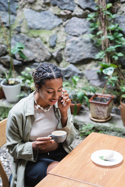 African american woman talking on smartphone and holding cup near dessert in outdoor cafe  - Zdjęcie, obraz
