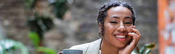 Smiling african american woman looking at camera near cup on cafe terrace, banner  - Foto, Imagen