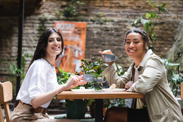 Smiling multiethnic girlfriends with coffee looking at camera on terrace of cafe  - Zdjęcie, obraz
