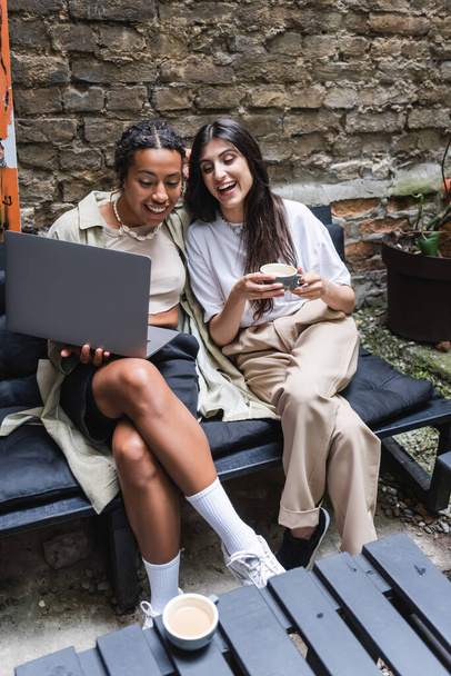 Cheerful interracial girlfriends with coffee using laptop in outdoor coffee  - Foto, Imagen
