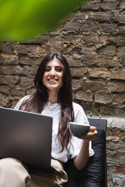 Cheerful brunette freelancer holding cup and laptop while looking at camera on terrace of cafe  - Foto, Bild
