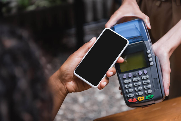 Cropped view of waitress holding payment terminal near african american client with smartphone in outdoor cafe  - Foto, Imagem