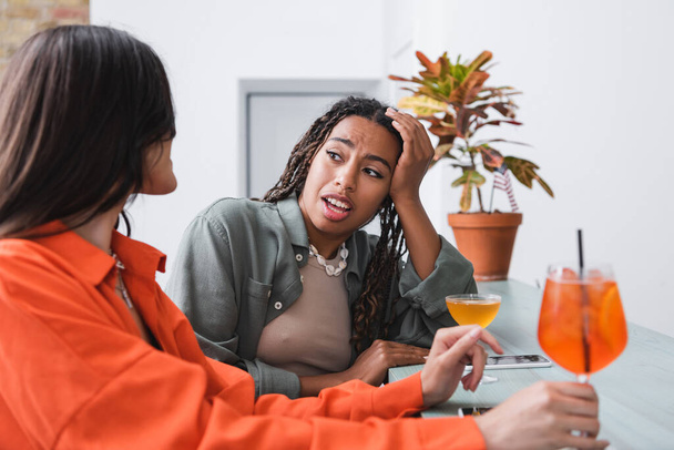 Африканская американка разговаривает с подругой возле коктейлей и смартфонов в кафе  - Фото, изображение