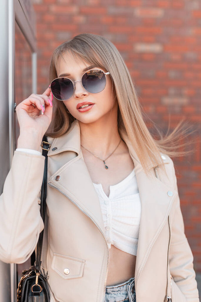 Fashionable young beautiful woman in casual fashion clothes with leather white jacket and bag wears a stylish round sunglasses, walks in the city near a brick wall - Zdjęcie, obraz