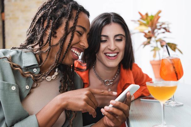 Αφροαμερικανή γυναίκα δείχνει το smartphone κοντά φίλη και κοκτέιλ στο καφέ  - Φωτογραφία, εικόνα
