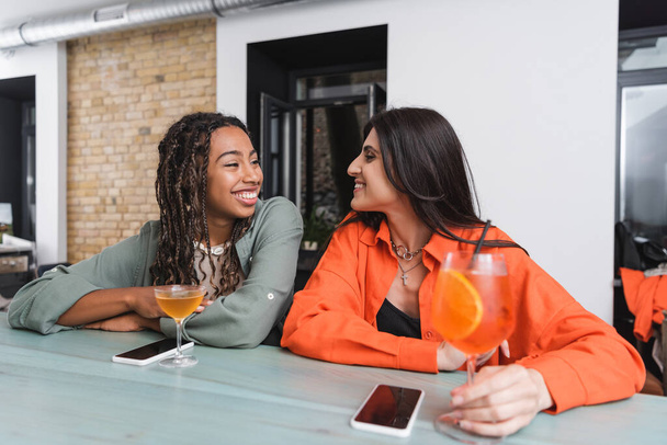 Мультиэтнические девушки разговаривают возле смартфонов и коктейлей в кафе  - Фото, изображение