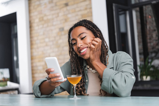 Позитивна афріканська жінка, яка користується мобільним телефоном біля коктейлю в кафе.  - Фото, зображення