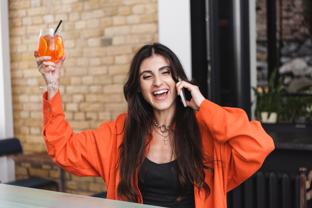Excited woman holding cocktail and talking on smartphone in cafe  - Photo, Image