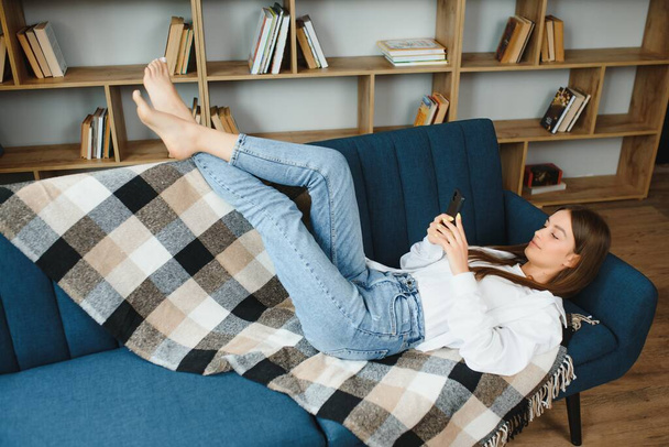 Young beautiful woman wearing white t-shirt on textile sofa at home. Attractive slim female in domestic situation, resting on couch in her lofty apartment. Background, copy space, close up - Photo, image