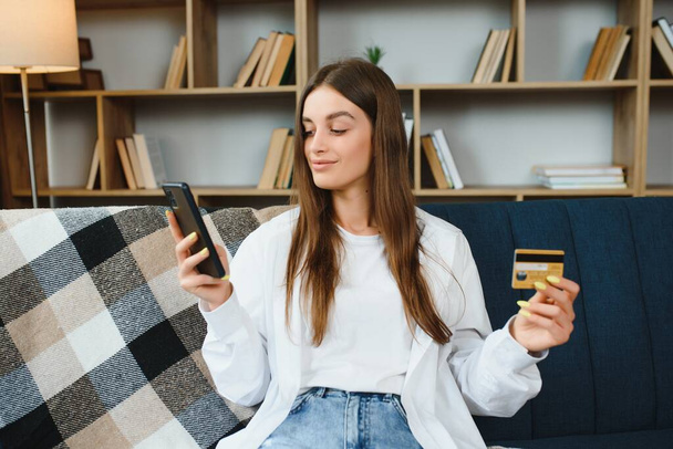 Happy young woman holding phone and credit card sitting on the couch. Online shopping - Φωτογραφία, εικόνα