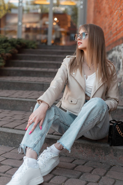 Модная красивая молодая хипстерша в солнечных очках в модной повседневной одежде в кожаной куртке, джинсах и белых кроссовках с сумочкой сидит на ступеньках за пределами торгового центра в городе - Фото, изображение