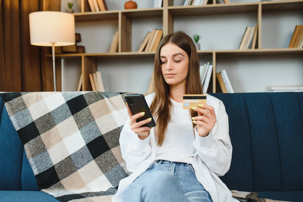 Happy young woman holding phone and credit card sitting on the couch. Online shopping - 写真・画像