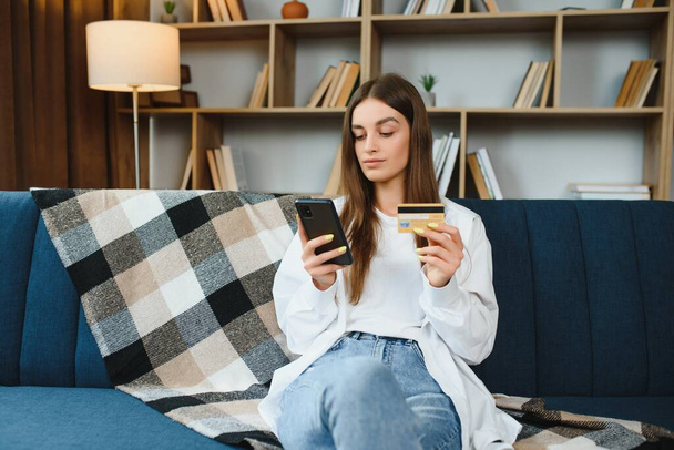 Happy young woman holding phone and credit card sitting on the couch. Online shopping - Foto, afbeelding