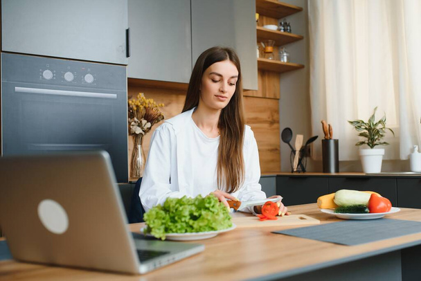 Joyeux jeune femme faire une salade à la cuisine, couper des légumes, regarder ordinateur portable
. - Photo, image