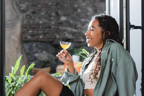 Side view of cheerful african american woman holding cocktail near window in cafe  - Foto, imagen