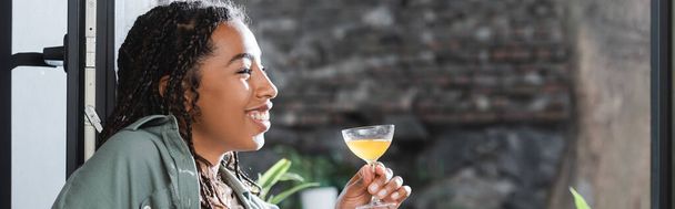 Side view of positive african american woman holding cocktail near window in cafe, banner  - Foto, afbeelding