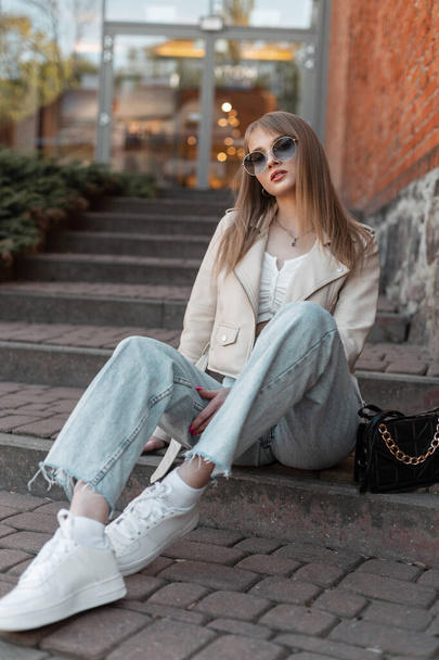 Beautiful fashionable hipster zoomer woman in fashionable street clothes with a white leather jacket, jeans and stylish sneakers with a purse is sitting on the steps in city near the shopping mall - Foto, Imagen