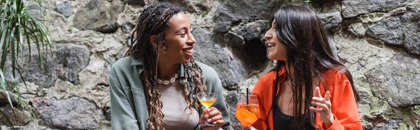 Positive multiethnic girlfriends holding cocktails while talking on terrace of cafe, banner  - Foto, Imagen