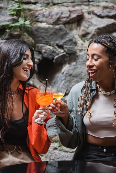 Positive multiethnic girlfriends clinking with cocktails on terrace of cafe  - Foto, Imagem
