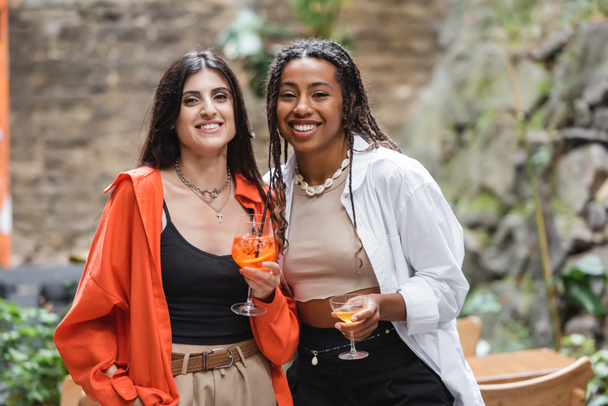 Positive multiethnic women holding cocktails and looking at camera on terrace of cafe  - Foto, Imagem