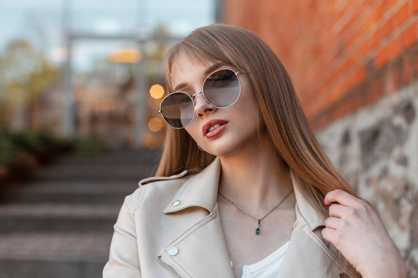 Urban female stylish portrait of a beautiful fashionable woman with vintage round hipster sunglasses in a leather jacket sits near a vintage mall on the street - Fotó, kép