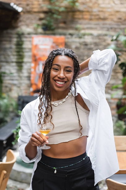 Cheerful african american woman holding cocktail on terrace of cafe  - Foto, imagen