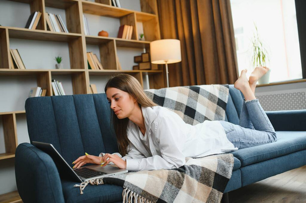 Freelancer working on laptop at home laying on comfortable beige sofa - Fotó, kép
