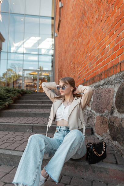 Trendy fashionable beauty girl model with fashion sunglasses in casual clothes with leather jacket, blue vintage jeans and purse sits and poses on the steps near a red brick mall - Фото, зображення