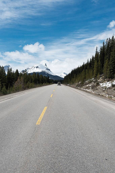 An empty road surrounded by pine trees with snow-capped mountains in the background in Banff, Canada - Photo, image