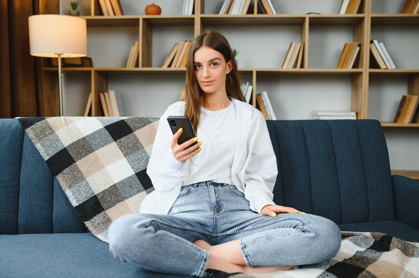 Smiling young Caucasian woman sit relax on couch in living room talk on video call. Happy millennial female rest on sofa at home have webcam digital virtual communication on cellphone - Fotografie, Obrázek