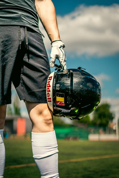 A vertical shot of a man holding with white nike gloves, a belgian Redbull american football helmet against his knees - Φωτογραφία, εικόνα