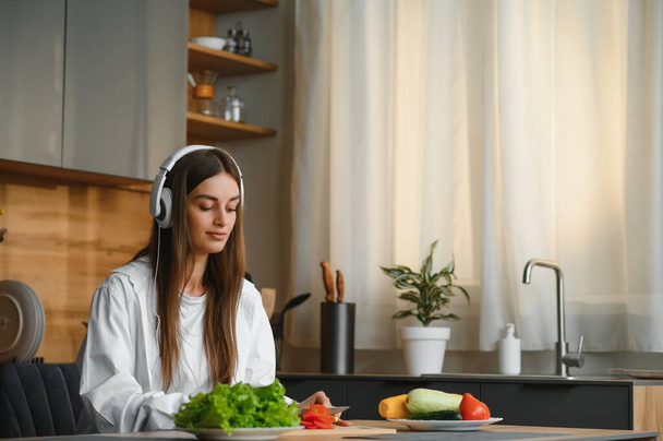 Adorable nice charming cheerful pretty beautiful stylish excited girl listening to music, singing in modern light white kitchen, cooking homemade dish with domestic products. - 写真・画像