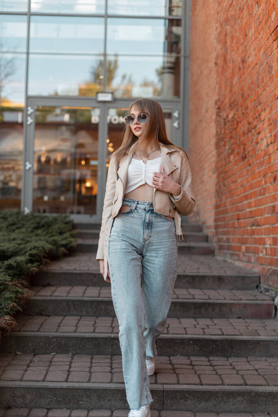 Stylish beautiful young woman with trendy sunglasses in a fashionable beige leather jacket with fashion vintage high waist jeans and white sneakers walks near the brick shopping mall - 写真・画像