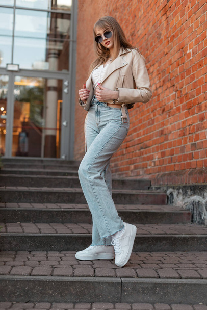 Beautiful young woman model hipster with trendy sunglasses in fashionable casual clothes with fashion leather jacket, high waist jeans and white sneakers walking near a shopping mall with brick wall - Φωτογραφία, εικόνα