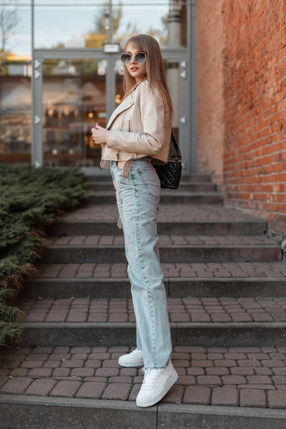 Stylish beautiful young woman with trendy sunglasses in a fashionable beige leather jacket with fashion vintage high waist jeans and white sneakers standing near the brick shopping mall - Valokuva, kuva