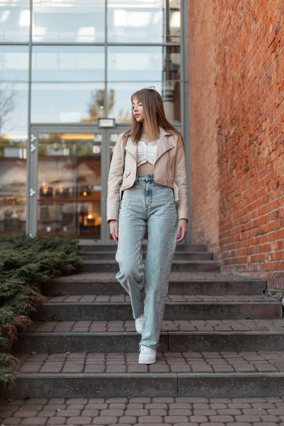 Trendy beautiful young woman in fashionable casual wear outfit with a leather jacket with bell-bottom high-waisted jeans and white sneakers walks near a red brick store building - Foto, Bild
