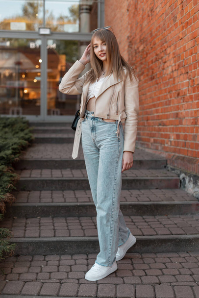 Happy beautiful young girl in fashionable clothes with a leather jacket, a vintage high-waisted jeans and white shoes walks down the steps in the city. Female urban casual style and fashion - Fotó, kép