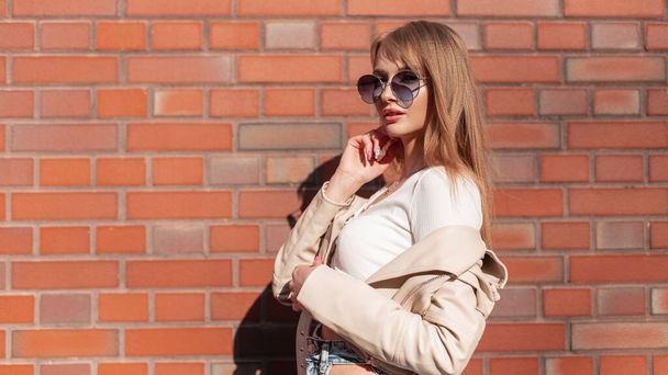 Fashionable beautiful stylish woman model hipster with round sunglasses in stylish casual clothes with leather jacket and top standing near the brick red wall - Foto, Imagen