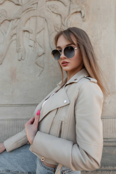 Fashionable young adorable girl model with stylish  sunglasses in fashion streetwear with leather beige rock jacket poses near a vintage wall on the street - Фото, изображение