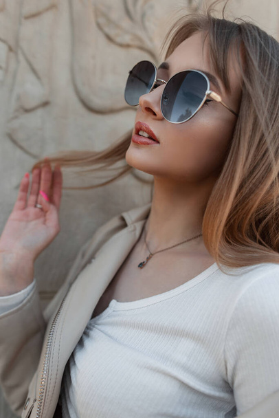 Fashionable portrait of a beautiful stylish pretty woman with a beautiful face with fashion sunglasses in trendy casual clothes with a top and a leather jacket on street near a vintage wall - Fotó, kép