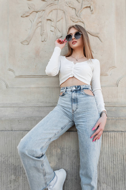 Stylish beautiful young girl model wears trendy sunglasses in a fashion white long-sleeved top with blue high-waisted jeans posing near a vintage wall. Urban female fashion and beauty - Фото, изображение