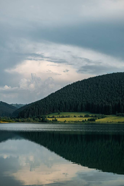 A vertical shot of a lake reflecting the forested mountain and the sky during sunset - Фото, зображення