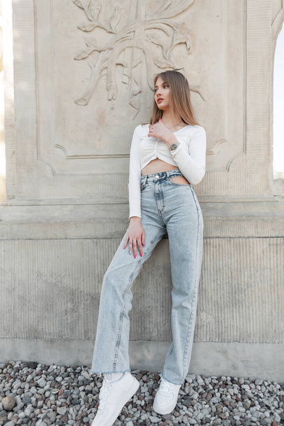 Fashion pretty woman hipster mode in trendy streetwear with stylish white long sleeve top, vintage height waisted jeans with white sneakers stands near a vintage column outdoors - Photo, image