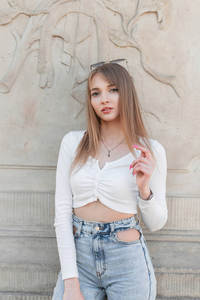 Beautifully young fashion girl in casual urban outfit with trendy white long sleeve top and blue fashionable high waist jeans stand and poses near a vintage wall outdoors and looks at the camera - Photo, image