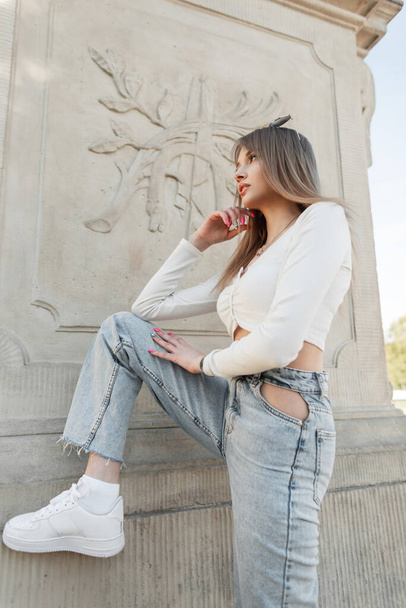 Fashionable beautiful girl with a slender body in fashion casual streetwear with white long sleeve top and blue high waisted jeans with white sneakers poses near a vintage column in the city - Photo, Image